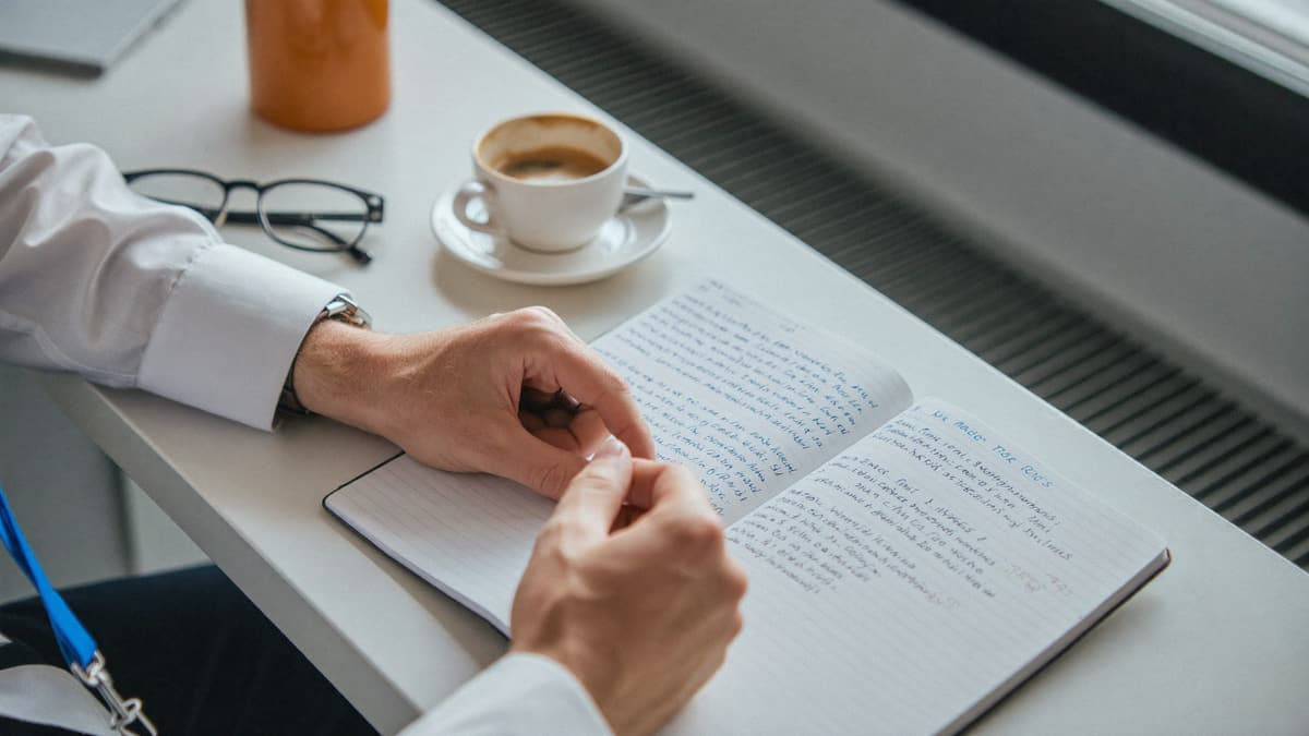Mains d'un praticien près d'un carnet de comptes manuscrit et d'un café, dans la lumière du matin